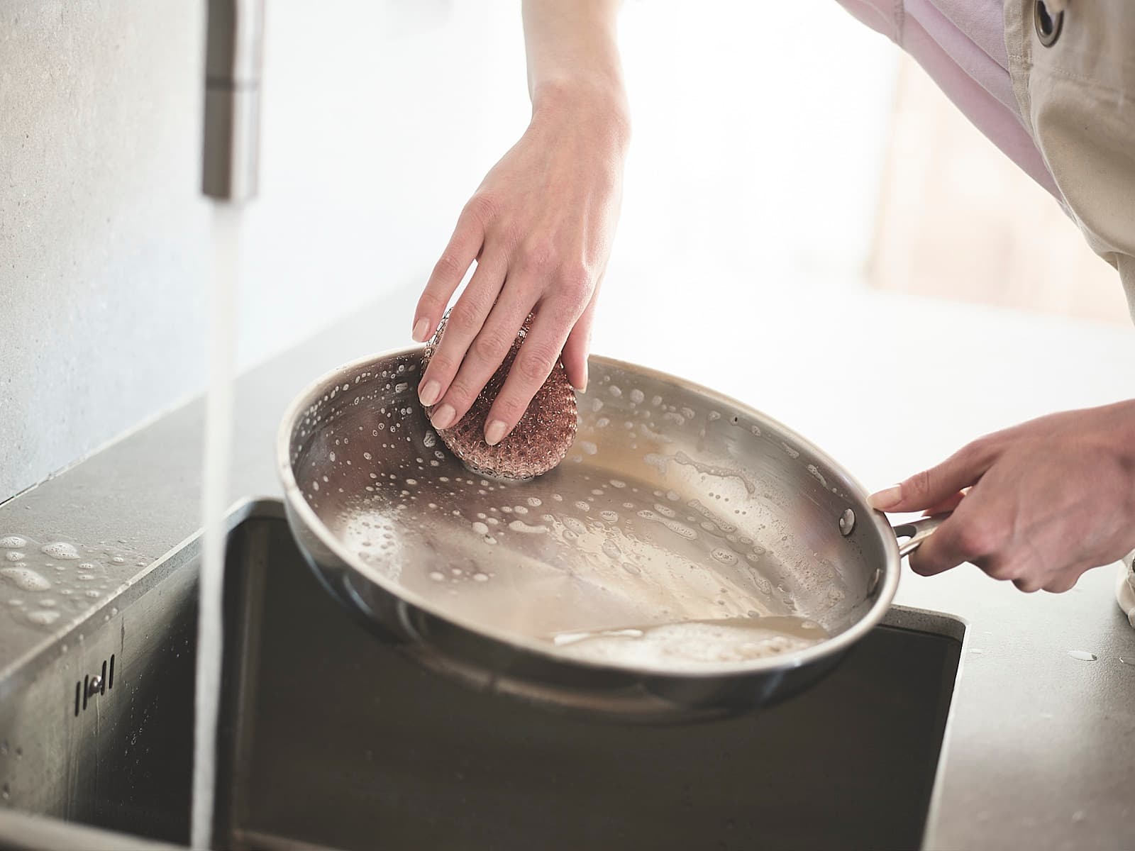 Scrubbing a stainless steel frying pan with a scouring pad under a running tap