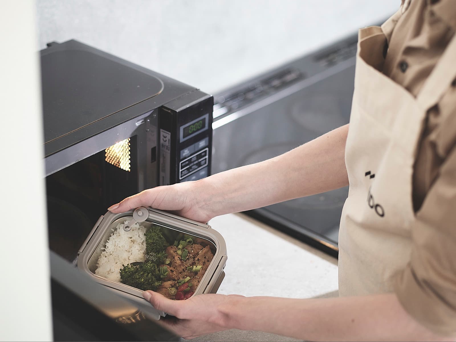 A SEIDO stainless steel lunch box of rice, broccoli and chicken being placed in a microwave