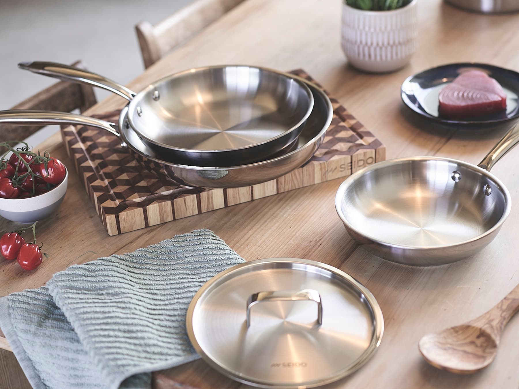 Three SEIDO stainless steel frying pans and a stainless steel lid on a wooden kitchen table