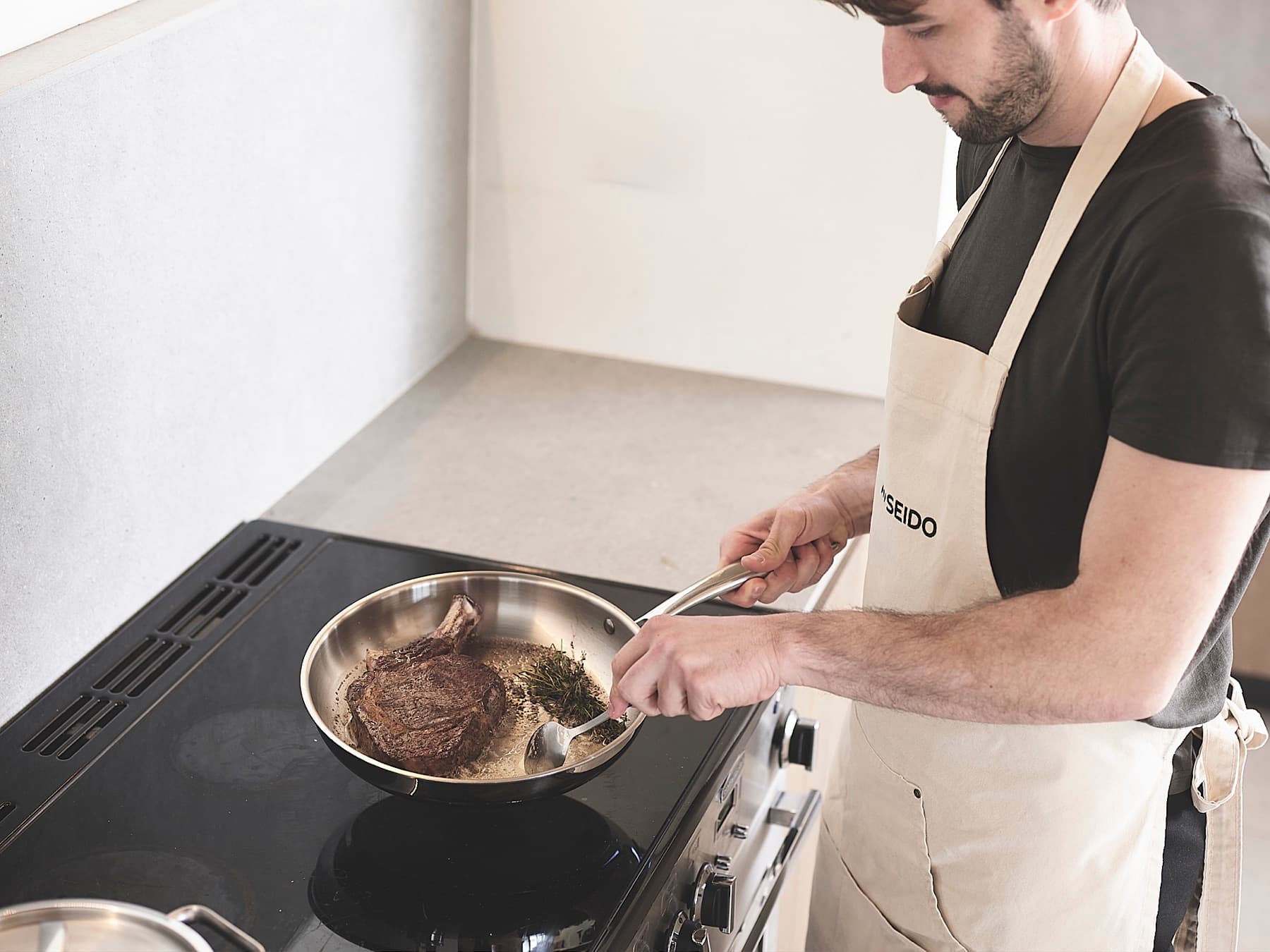 Cook spooning butter over a bone-in steak with herbs in an uncoated stainless steel frying pan
