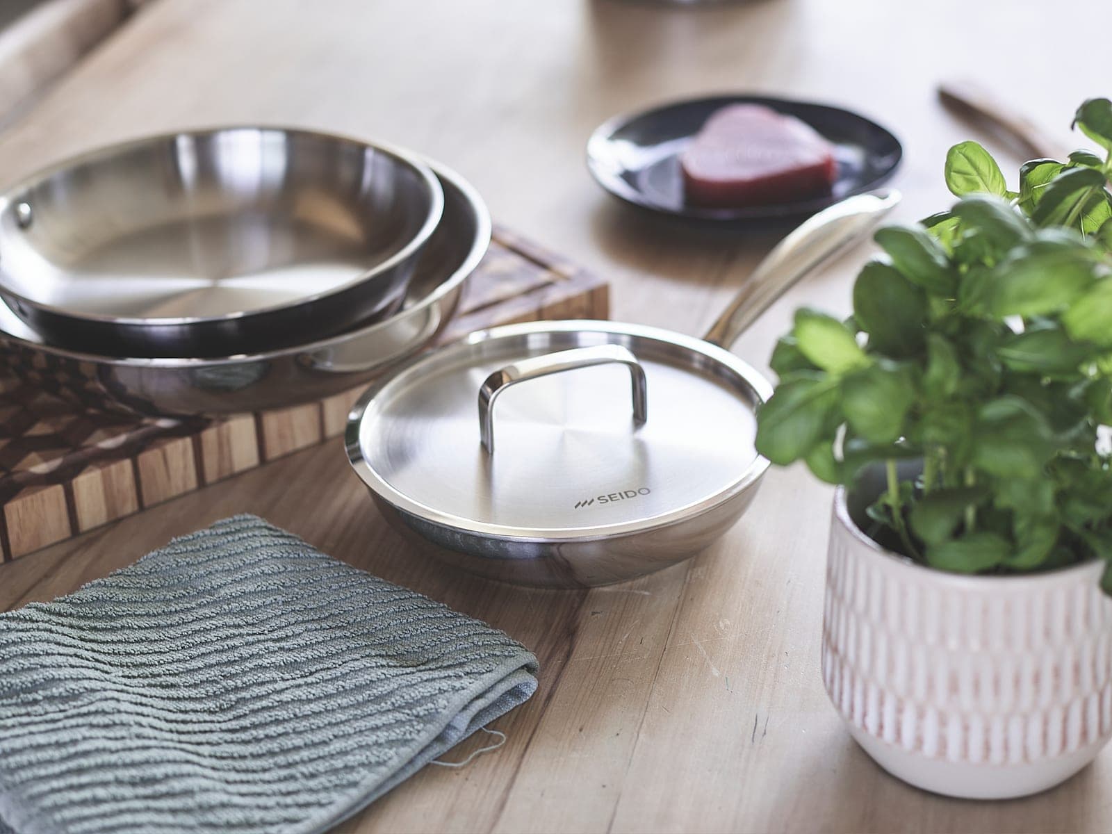 Stainless steel frying pans with their lid on a kitchen table beside steak and basil