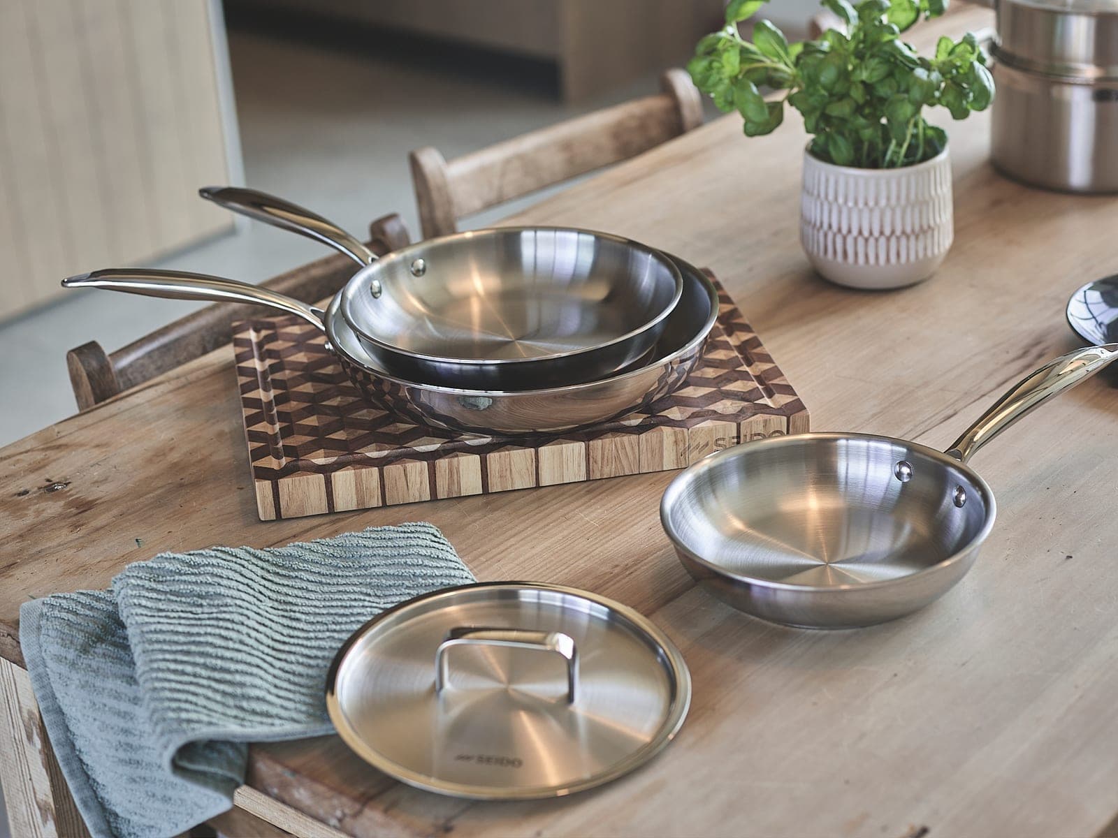 Three sizes of SEIDO stainless steel frying pan and a matching stainless steel lid on a wooden kitchen table